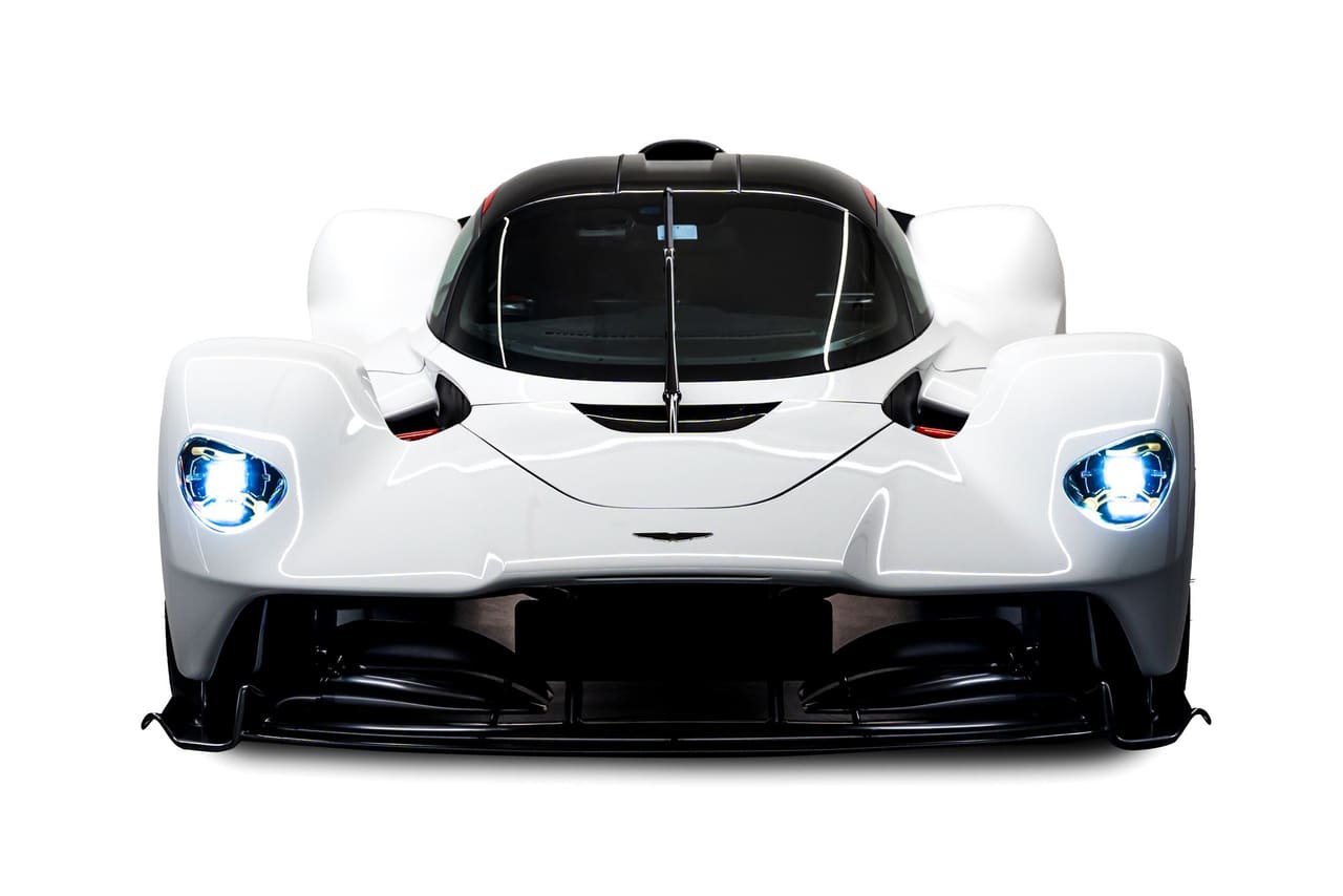A white Aston Martin Valkyrie head-on, headlamps lit, with the cockpit in the centre and open channels on either side of it beneath the bodywork