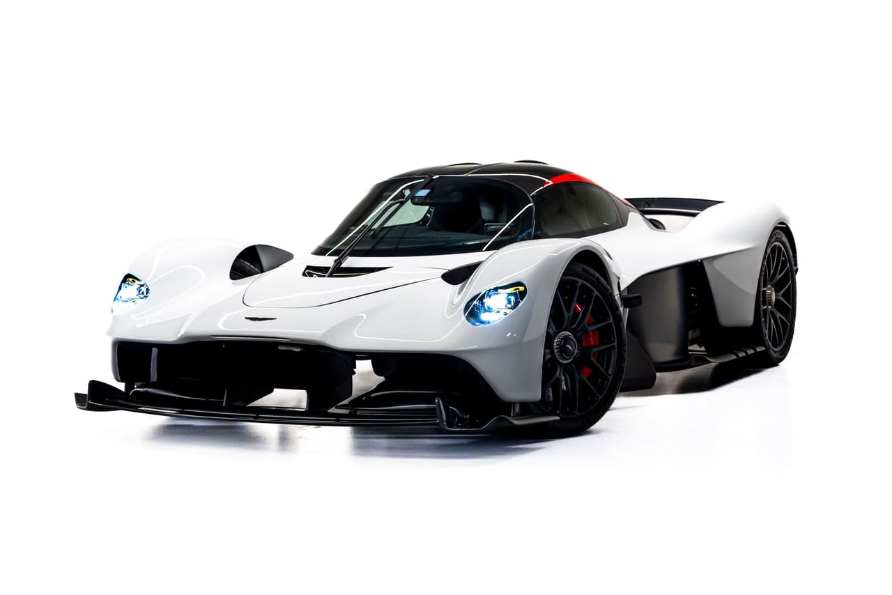 A white Aston Martin Valkyrie from the front three-quarter on a white studio floor, headlamps lit, with its long front splitter, black wheels and red calipers