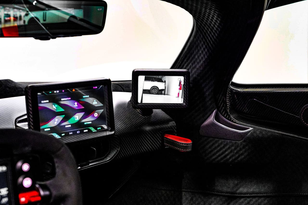 Inside an Aston Martin Valkyrie: a screen at the base of the windscreen pillar showing the rear view, beside the carbon fibre door frame and the central display