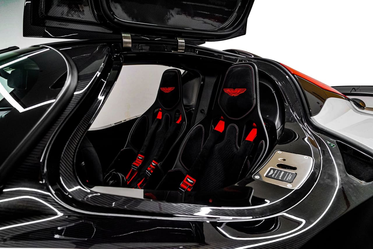 The two seats of an Aston Martin Valkyrie through the open door, finished in black with red harnesses and red-trimmed headrests, set into the exposed carbon fibre structure