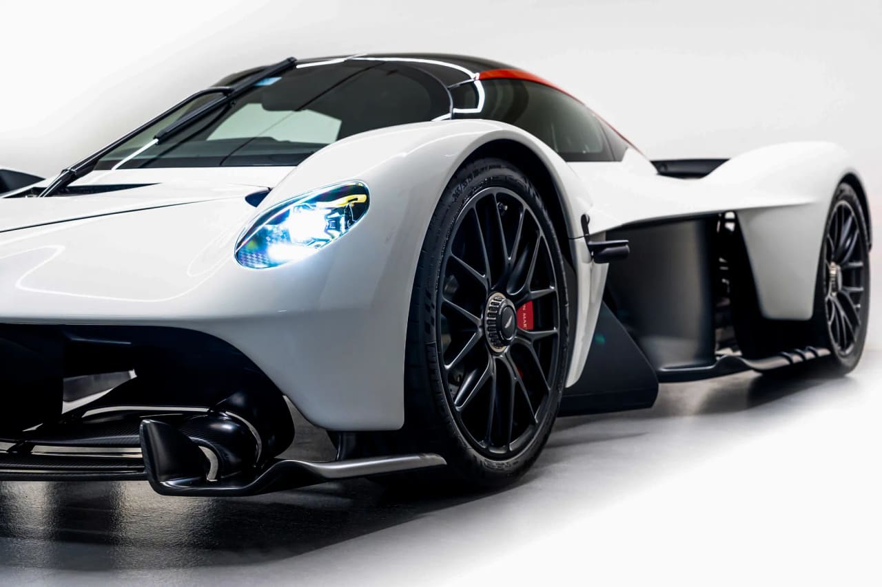 A white Aston Martin Valkyrie seen low from the front three-quarter, headlamps lit, with black wheels, red brake calipers and a red stripe over the roof